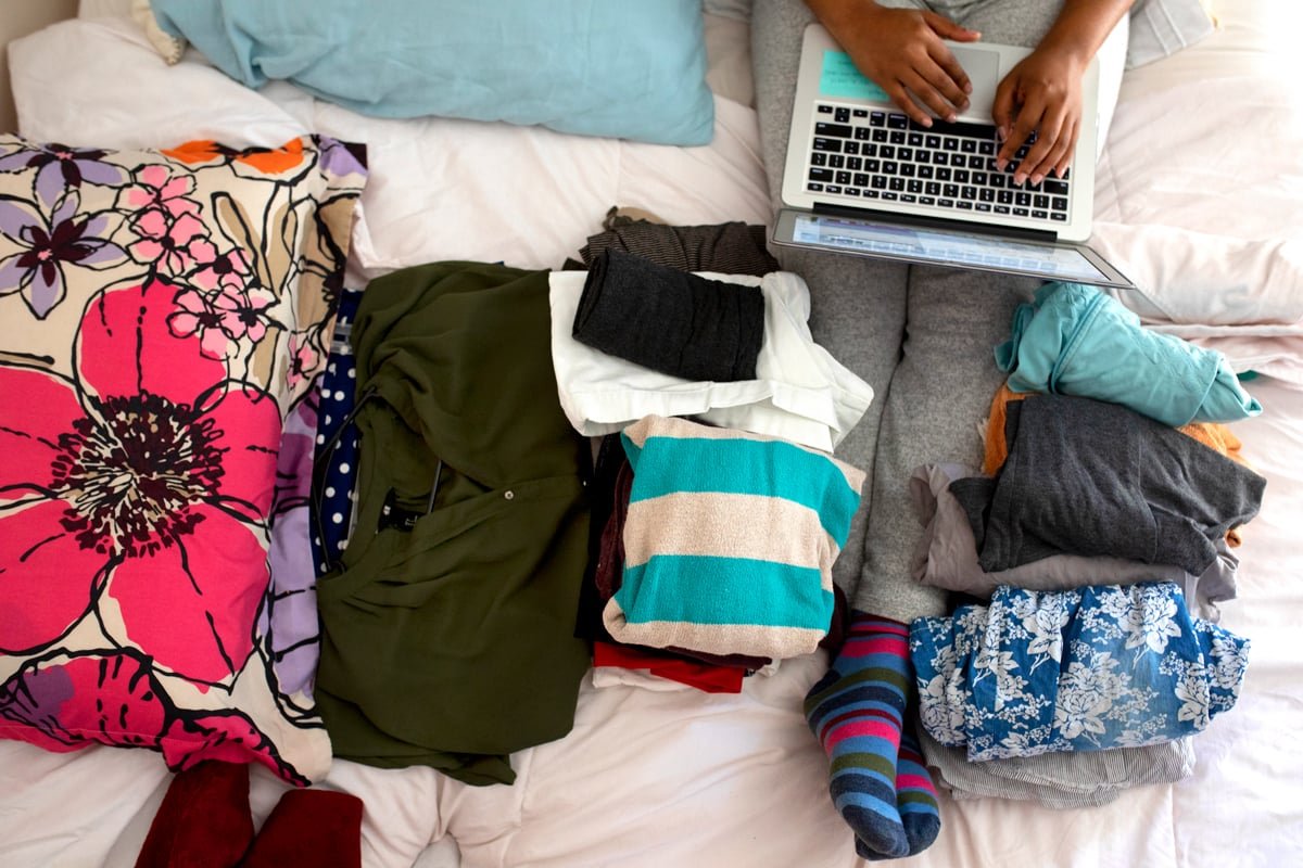 A woman types on a computer with clothes surrounding her that she's trying to sell online. 