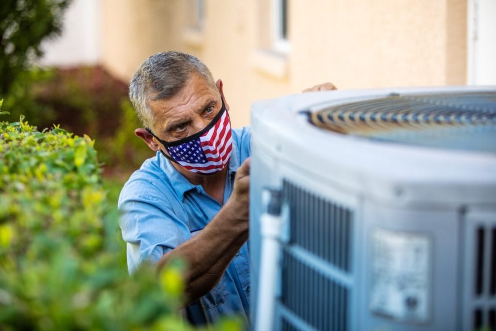 A man works on an HVAC system outside while wearing a face mask decorated with the American flag. 